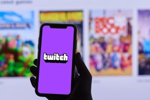 A phone with the Twitch app open is held before a blurred monitor screen.