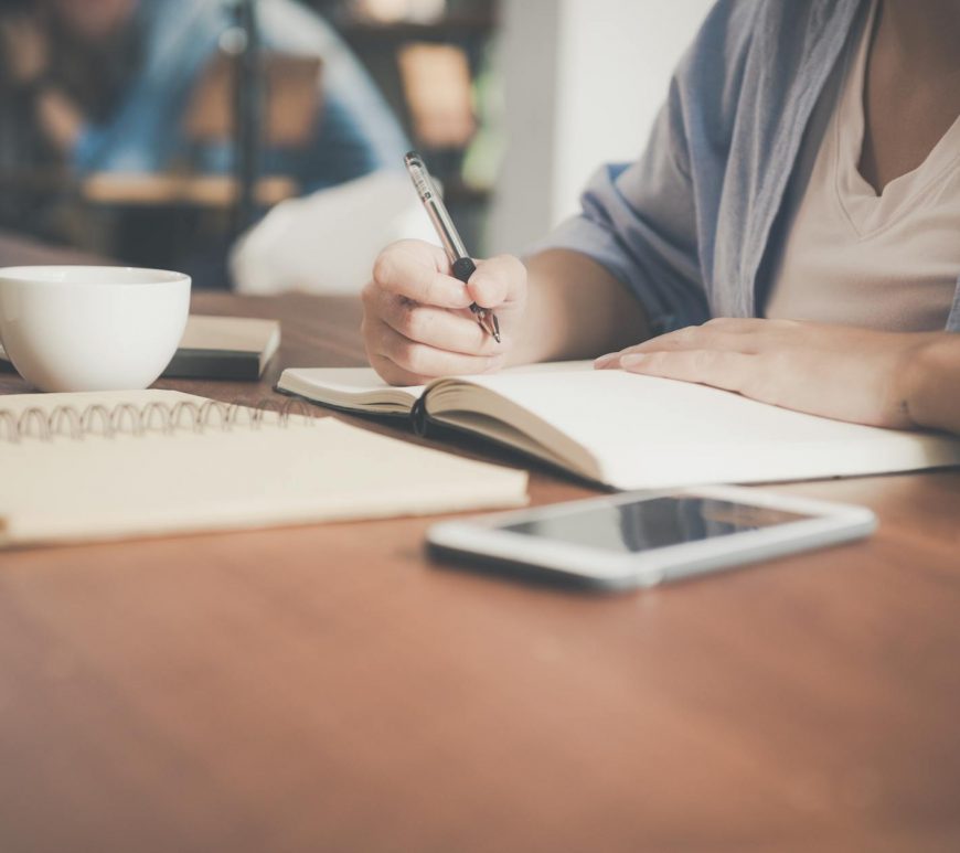 woman writing on a notebook beside teacup and tablet computer