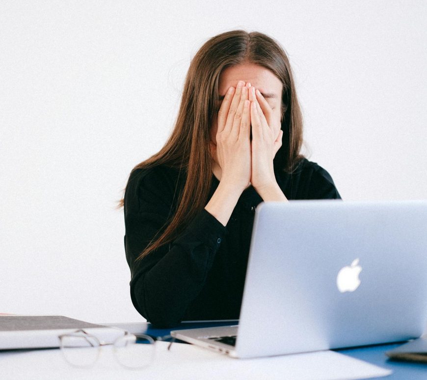 A person hiding their face with their hands in front of a Macbook.