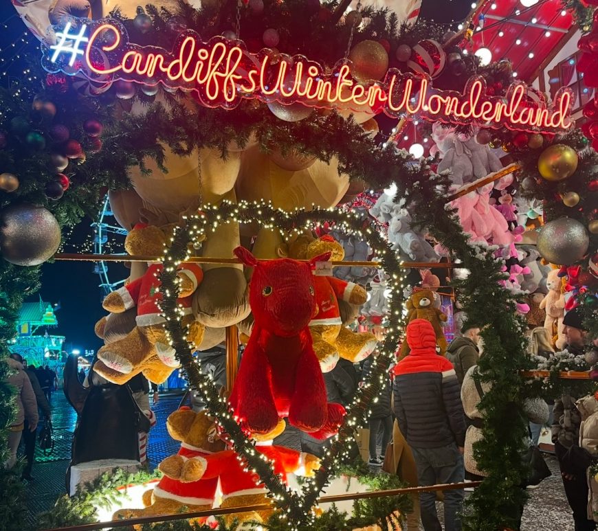 Winter Wonderland Sign with stalls and lights in the background with a Welsh Dragon teddy in the centre