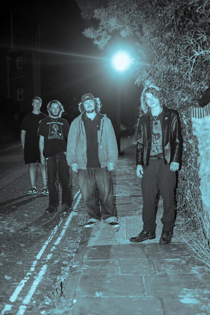 Four band members stood in a line of the pavement with a street lamp shining behind them